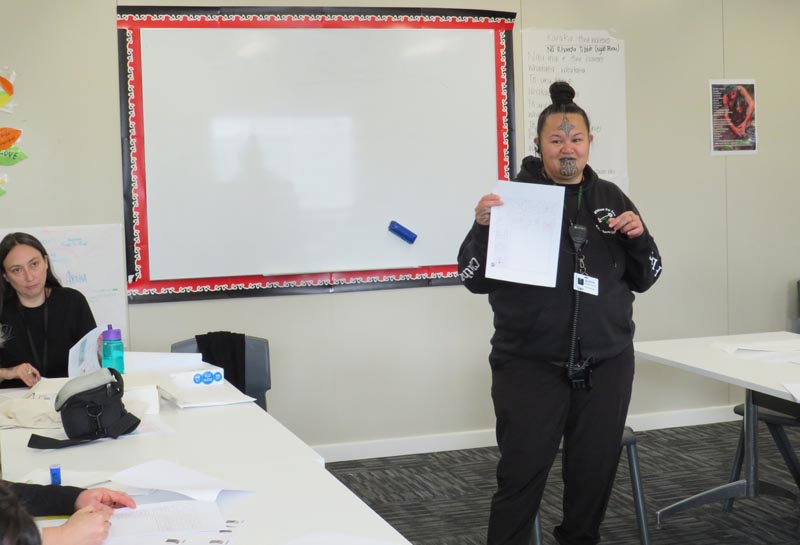 Christine Harvey stands in front of a whiteboard, holding up a piece of A4 paper. In the lefthand corner of the image is Tōmairangi Taiepa 
