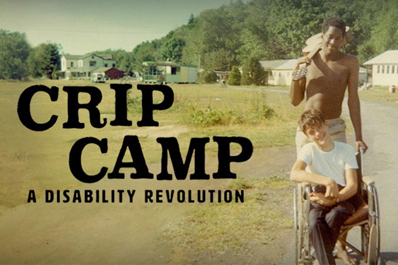 Cover image for Crip Camp film in which a man carries a guitar over one shoulder while pushing another man in a wheelchair.
