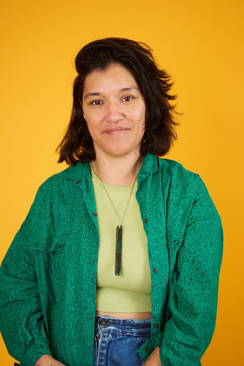 A portrait image of Pelenakeke Brown, wearing a green jacket and posed in front of a yellow background