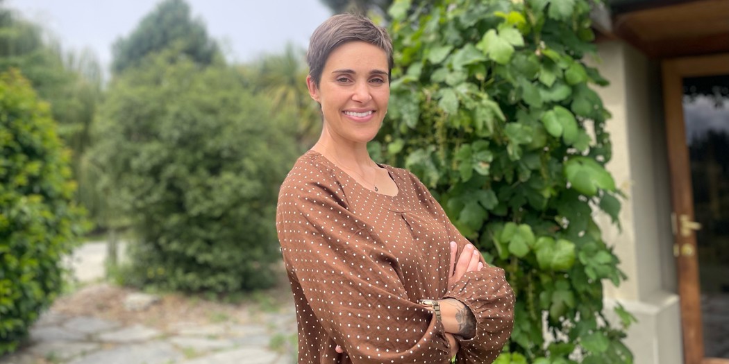 Victoria wears a brown polkadot top and smiles broadly while standing in front of green foliage.