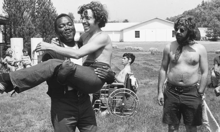 A still from Crip Camp in which a man holds another in his arms in a field while others sit in the background in wheelchairs and chairs.