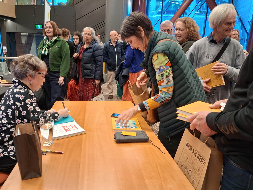 Trish Harris seated at a table, signing her book at the book launch. She is surrounded by people, many wanting copies of the book signed.