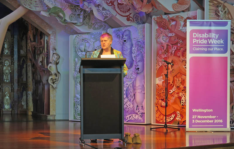 Robyn Hunt stands at a wooden podium addressing a large audience at Disability Pride Week 2016