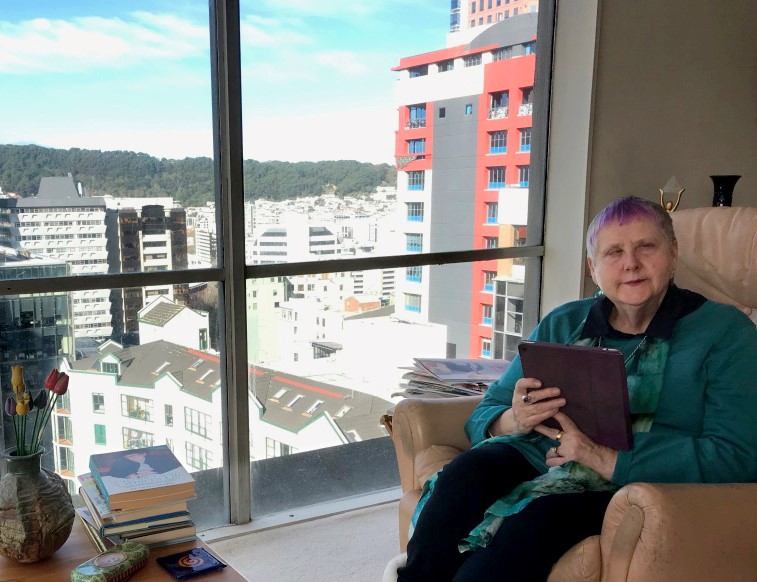 Robyn Hunt sits in front of a large window looking out to Wellington city with a book in her hands.